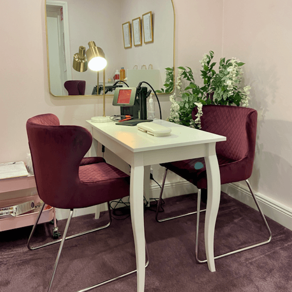 permanent/welded jewellery studio with two purple chairs opposite sides of a white desk with a small welding machine and mirror hanging on a pink wall