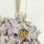 a Close Up Picture of a gold dara knot strength necklace with purple flowers in the background