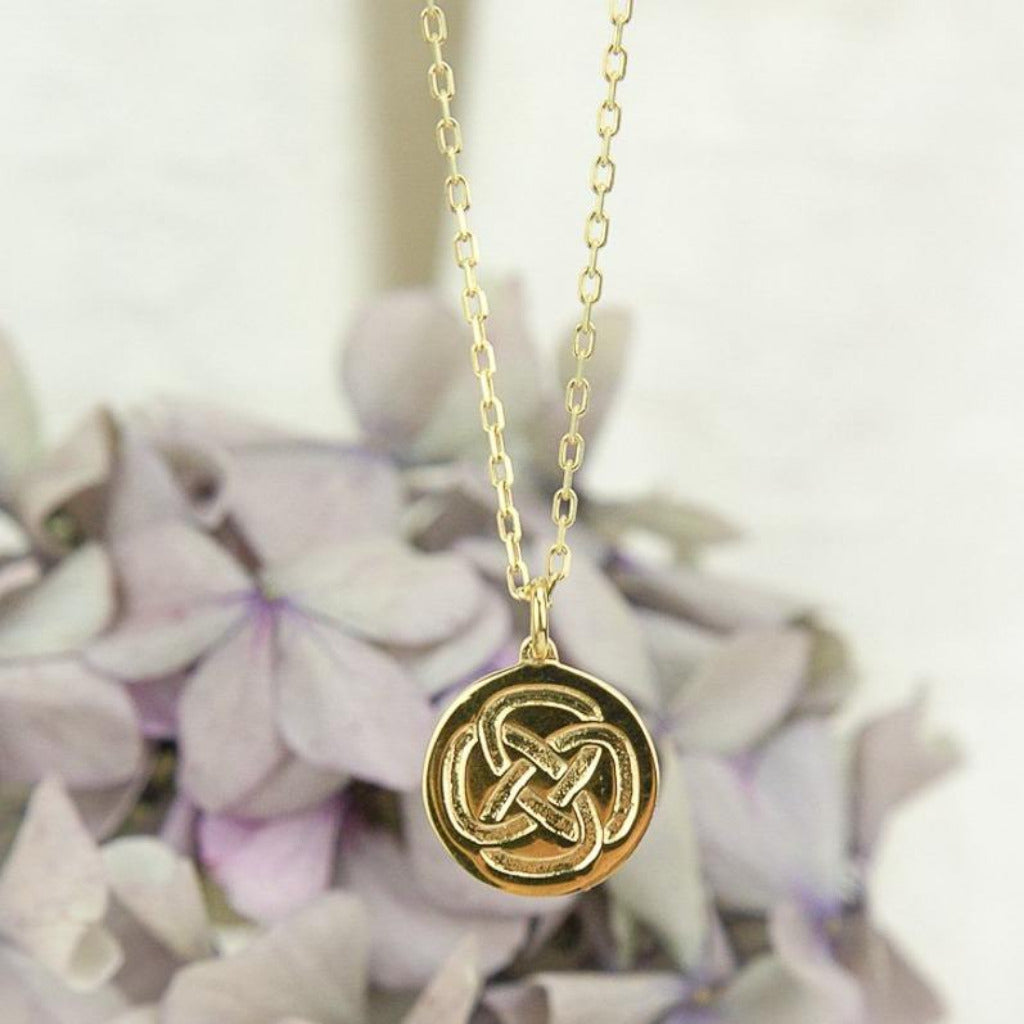 a Close Up Picture of a gold dara knot strength necklace with purple flowers in the background